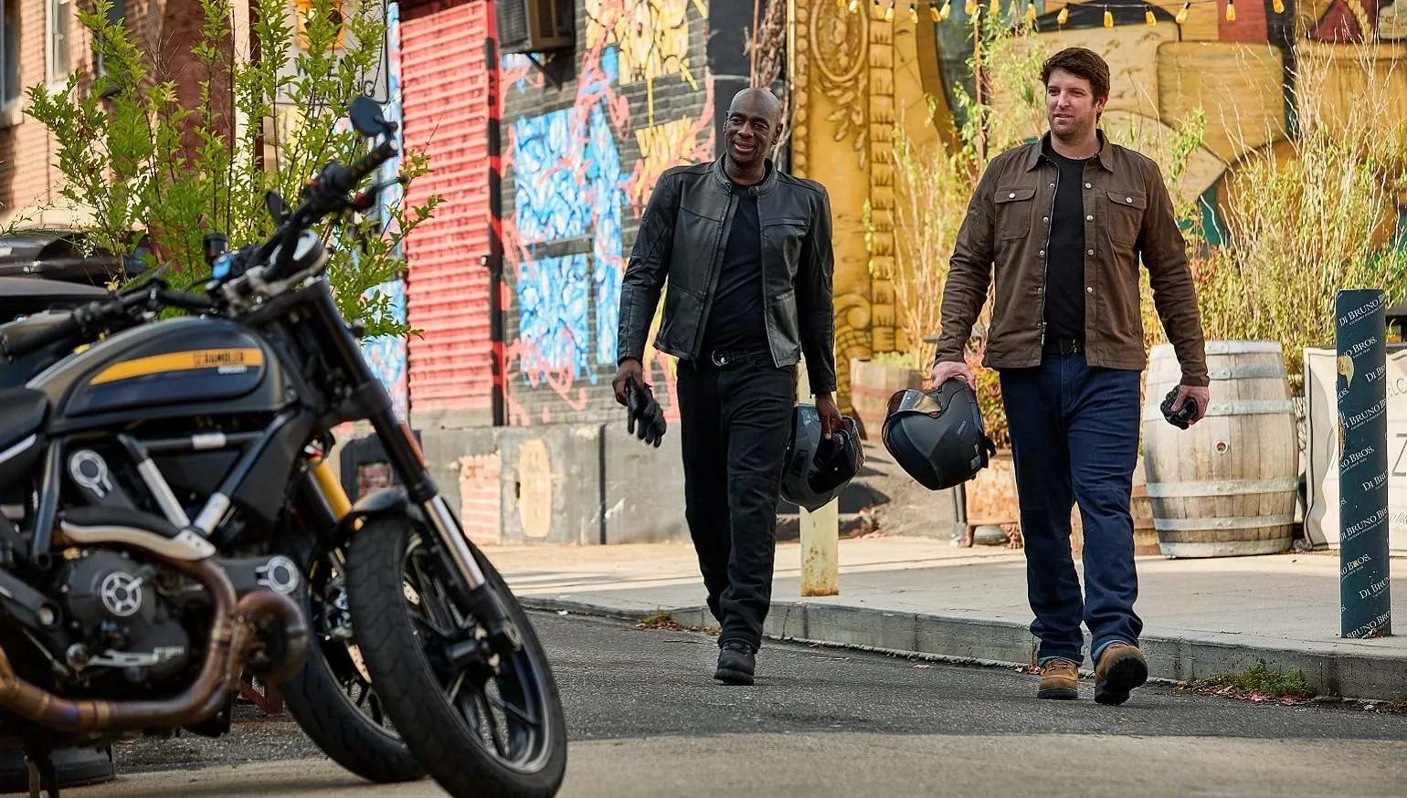 Two men wearing jackets walk together on a city street after parking their motorcycles.