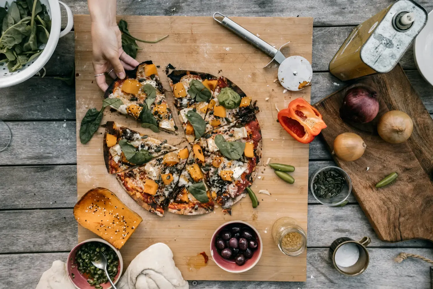 A hand reaches for a slice of homemade pizza topped with spinach and squash, surrounded by fresh ingredients and kitchen tools on a rustic wooden table.
