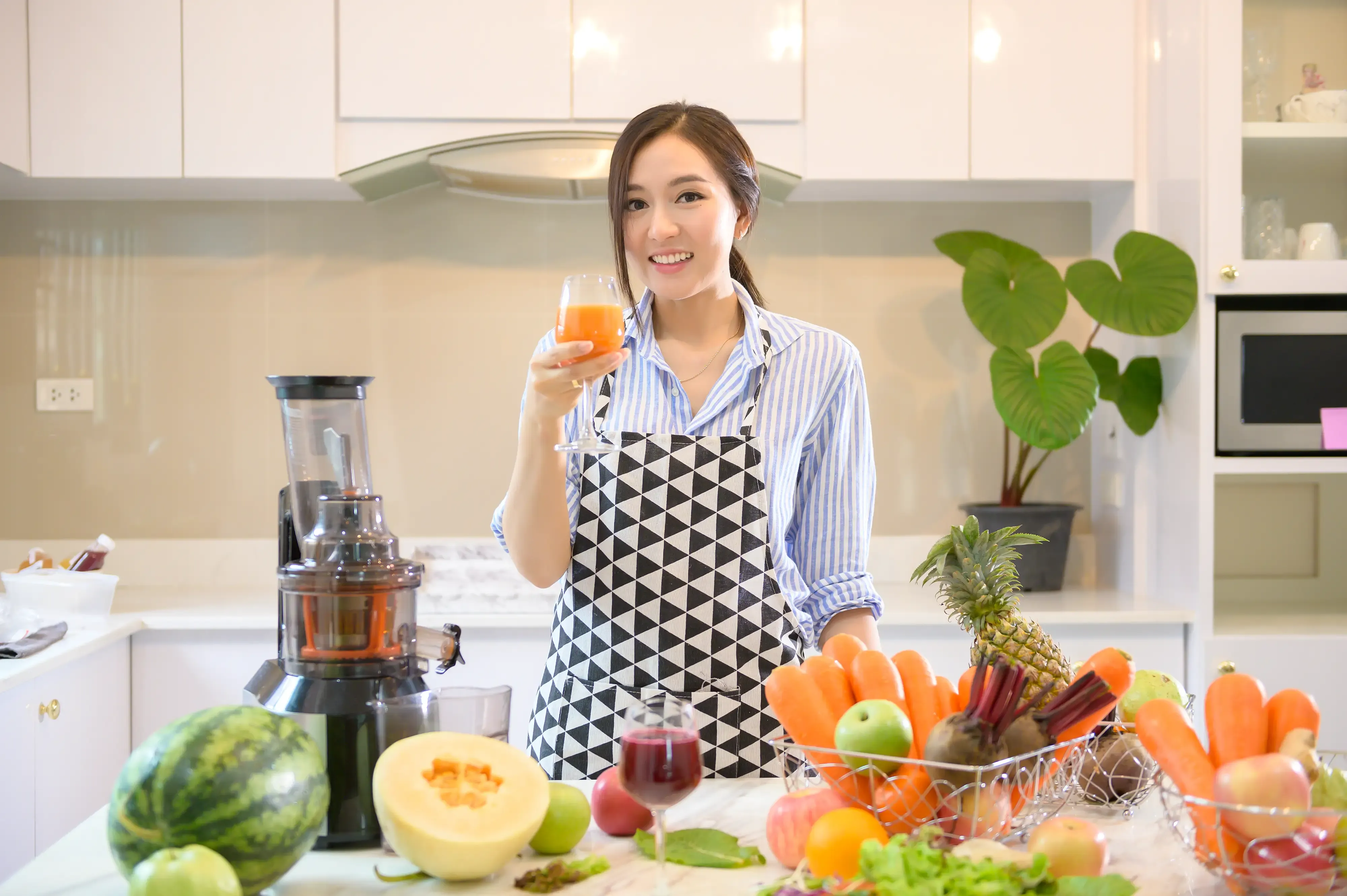 A woman holding a glass of juice standing behind a kitchen counter full of fruits on top and a juicer