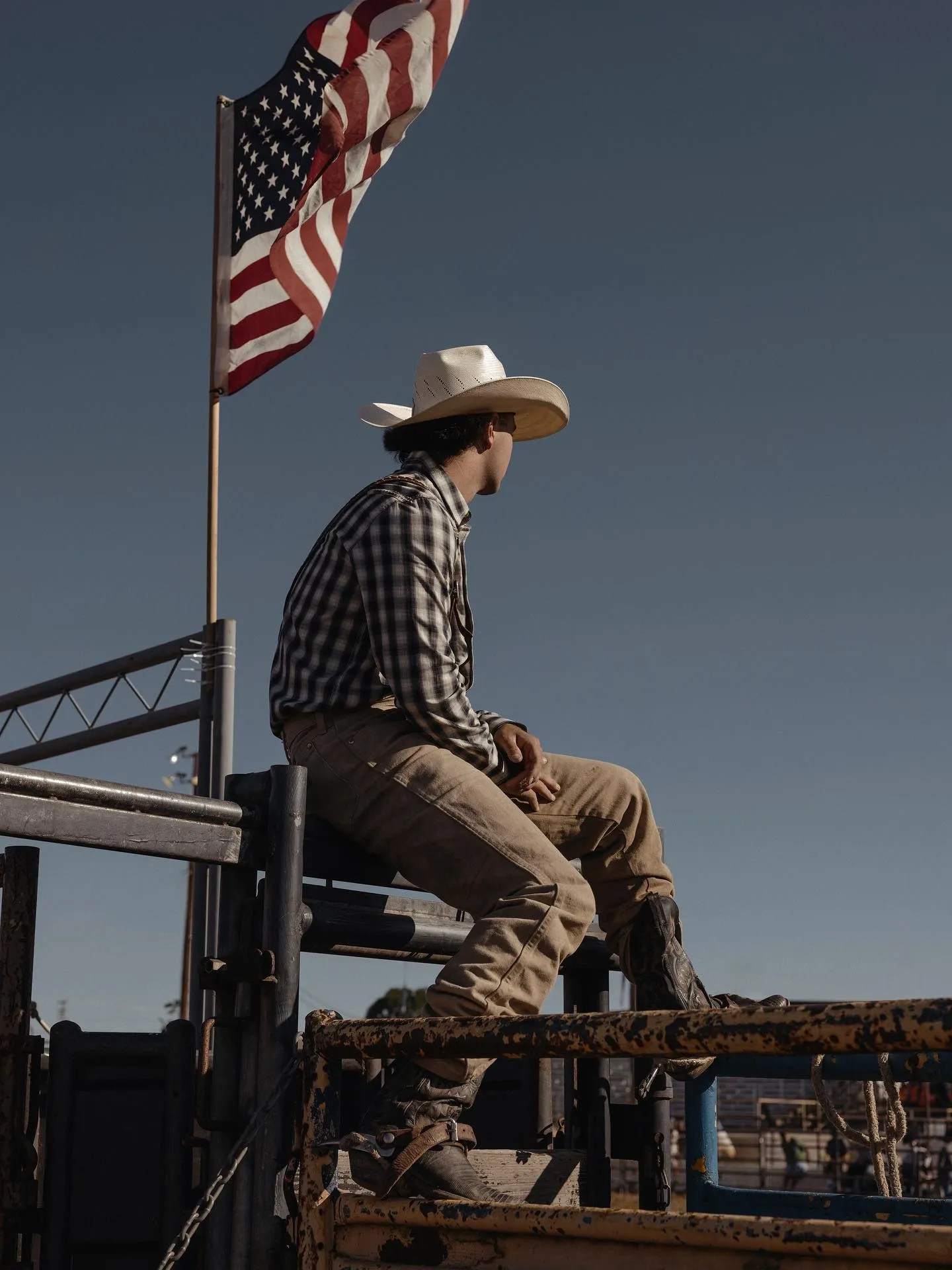 A cowboy wearing a hat and boots sits on a fence with an American flag flying above him.