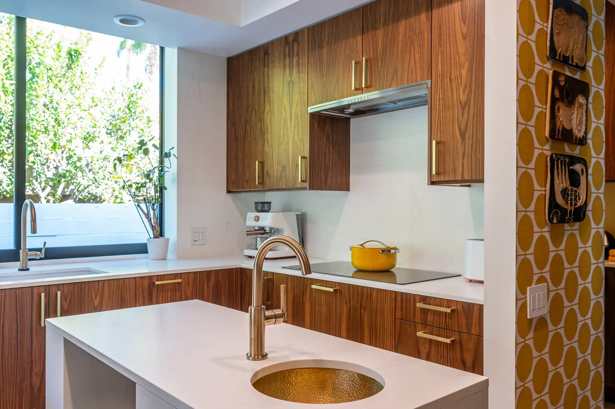 A modern kitchen features rich, wood-grain cabinets with brass hardware, a white countertop, and a kitchen island with a unique, circular brass sink.