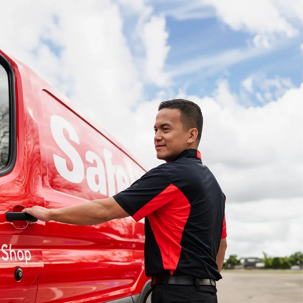 A Safelite technician in a black and red uniform is opening the door of a red service van.